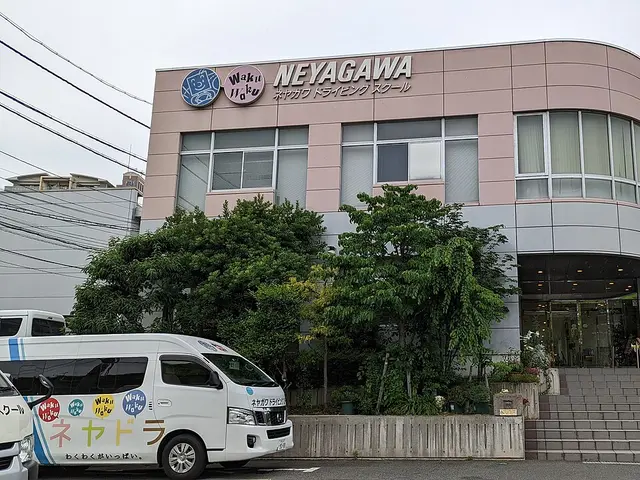 The pink and grey exterior of Neyagawa Driving School in Japan, with a branded minibus parked outside.