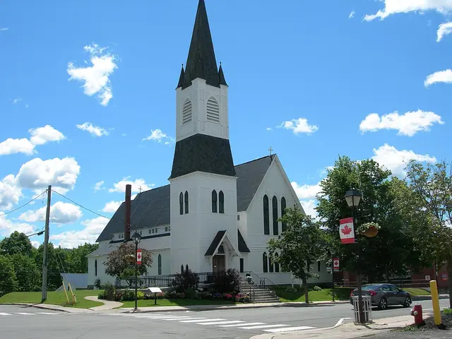 St. Luke's Anglican Church in Woodstock, New Brunswick, a white wooden church with a tall black spire, seen from the street corner with Canadian flag banners.