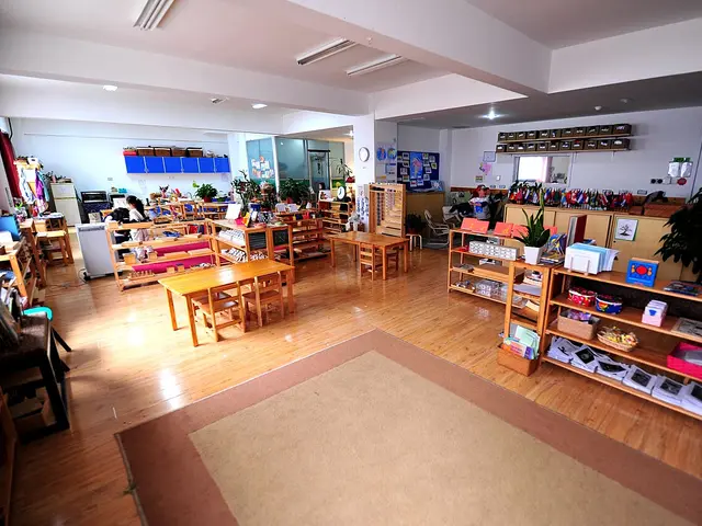 A Montessori classroom with wooden shelves of learning materials, small tables and chairs, and a carpeted area.